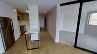 This contemporary single-floor residence showcases a thoughtfully designed open-concept layout that seamlessly connects the kitchen and living areas. The kitchen is a standout feature, boasting warm wood cabinetry, a clean white countertop island with minimalist black metal legs, and stainless steel appliances, creating a stylish and functional cooking space. Light hardwood flooring extends throughout the main living areas, lending warmth and visual continuity to the home. A long, well-lit corridor connects the living spaces to additional rooms, offering a sense of depth and flow throughout the residence. The living area transitions to carpeted flooring, providing a comfortable contrast to the hardwood. Crisp white walls and clean architectural lines give the space a bright, modern aesthetic, while recessed and track lighting ensures well-distributed illumination. The overall ambiance is sleek and contemporary, with a neutral palette that offers a versatile canvas for personalization. The open layout and cohesive design make this space feel both spacious and inviting, ideal for modern living.