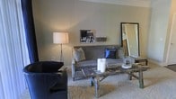 This well-appointed single-floor space showcases a refined sense of style through its carefully chosen furnishings and cohesive design aesthetic. The living area serves as the heart of the space, centered around a plush gray upholstered sofa accented with decorative pillows, including striking royal blue velvet cushions that add a bold pop of color against the neutral palette. A rustic, distressed wood coffee table sits at the center, providing an earthy contrast to the more polished elements of the room and lending a touch of character to the overall design. Complementing the seating arrangement is a dark leather accent chair, adding depth and variety to the furniture selection. The walls are finished in soft, muted gray-green tones that contribute to the calm and sophisticated atmosphere throughout. A large floor-length mirror leans against one wall, enhancing the sense of openness and light within the space. Subtle decorative touches — including pillar candles, a framed black-and-white photograph, and a tall floor lamp — complete the room's curated look. Plush wall-to-wall carpeting underfoot adds warmth and comfort, tying together all the design elements into a cohesive, welcoming environment ideal for relaxed living.