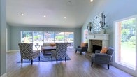This single-floor space exudes a warm, welcoming ambiance defined by its vaulted ceilings and an abundance of natural light. The main living area is generously proportioned, centered around a classic stone fireplace with a crisp white mantle, flanked by decorative wall sconces and framed artwork. The fireplace serves as a striking focal point, complemented by a curated seating arrangement featuring patterned accent chairs in bold geometric prints, grey upholstered chairs with vibrant orange throw pillows, and a neutral sofa anchored by a blue area rug. Light hardwood floors run throughout, lending warmth and continuity to the space. Large, panoramic windows line the far wall, framing picturesque views of a lush, landscaped outdoor area with mature trees, flowering plants, and what appears to be a patio or courtyard. The seamless connection to the outdoors floods the interior with natural light and creates an open, airy atmosphere. Recessed lighting in the vaulted ceiling further enhances the brightness of the room. The overall aesthetic is transitional, blending classic architectural elements with contemporary furnishings to create a comfortable and visually dynamic living environment.