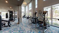 This state-of-the-art fitness center boasts a thoughtfully designed layout optimized for a variety of exercise routines. The space features high ceilings and large windows, allowing abundant natural light to flood the area, creating an open and energizing atmosphere. The flooring throughout is a distinctive mosaic-style carpet in shades of blue, gray, and teal, adding a vibrant touch to the environment.

The center is divided into multiple zones, each dedicated to specific workout types. A comprehensive weight training area includes a range of resistance machines and free weights, with ample space for strength exercises. The cardio section features modern equipment such as treadmills, ellipticals, and stationary bikes, strategically placed to offer users pleasant outdoor views while exercising.

Additional amenities include a functional training area with kettlebells and a wall-mounted TV for guided workouts. The facility also incorporates a beverage cooler for hydration and convenient storage options. Safety features like ceiling-mounted sprinklers and emergency exit signs are discreetly integrated into the design.

The overall aesthetic combines practicality with contemporary style, utilizing a neutral color palette accented by the vibrant flooring. Structural elements like exposed beams and columns are incorporated seamlessly, contributing to the spacious feel of the gym. This well-appointed fitness center provides a comprehensive and inviting space for users to pursue their health and wellness goals.