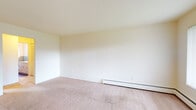This single-floor unit offers a straightforward, functional layout with distinct living, sleeping, kitchen, and bathroom areas. The living room is generously sized, featuring neutral wall-to-wall carpeting, crisp white walls, and a baseboard heating system that runs along the exterior wall. Large windows with vertical blinds allow natural light to fill the space, creating a bright and open atmosphere. A secondary carpeted area, accessible from the main living room, provides additional flexible space, also equipped with baseboard heating.

The galley-style kitchen is efficiently designed, featuring warm wood cabinetry above and below white laminate countertops, a stainless steel sink, and hardwood-style flooring. Black appliances, including a stove and oven, complete the kitchen's functional setup.

The bedroom offers comfortable proportions with plush carpeting, a window for natural light, and a full-width louvered bifold closet, providing ample storage space.

The bathroom features hardwood-style flooring, a white vanity with drawers, a wall-mounted mirror, and a tiled tub and shower surround in a muted blue-grey palette, adding a subtle color accent to the otherwise neutral interior. A window provides natural ventilation to the space.