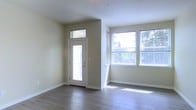 This single-floor space offers a clean, contemporary design characterized by neutral beige walls, white trim, and smooth hardwood-style flooring that runs throughout the interior. The entryway is defined by a white paneled door featuring a full-length glass insert and a transom window above, allowing natural light to filter in from the moment you step inside. Adjacent to the entry, a dedicated windowed nook provides an abundance of natural light through a wide set of double-hung windows fitted with blinds, offering views of surrounding greenery outdoors. Sunlight casts bright, natural patterns across the floors, enhancing the airy and open ambiance of the space. The layout is open and unobstructed, making it highly versatile for a variety of furnishing configurations. White baseboards and clean wall finishes contribute to the overall sense of brightness and modernity. The single-floor design ensures seamless flow throughout, while the generous window placement ensures consistent daylighting across the interior. Overall, the space presents a fresh, minimalist aesthetic well-suited for comfortable, everyday living.