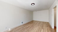 This single-floor unit offers a clean, minimalist aesthetic defined by neutral white walls and light wood-tone laminate flooring that runs consistently throughout the space. The main living area is a well-proportioned, open room illuminated by a flush-mount ceiling light fixture. A wall-mounted heating unit and multiple electrical outlets are conveniently positioned along the baseboards, adding practical functionality to the space.

The kitchen is efficiently laid out, featuring white cabinetry with a granite-style countertop and an undermount sink. A window above the sink frames a pleasant outdoor view of mature trees, allowing natural light to brighten the space. Black appliances, including an electric range and a full-size stainless steel refrigerator, complete the kitchen's functional setup.

The bathroom is finished with a glass-enclosed walk-in shower featuring built-in shelving, chrome fixtures, and a clean white surround. A vanity with a granite-style countertop and white cabinetry mirrors the design language found throughout the unit.

An additional room, accessible via a paneled door, provides a versatile space suitable for use as a bedroom or office. The consistent flooring, neutral palette, and practical finishes give the unit a cohesive, move-in-ready character.