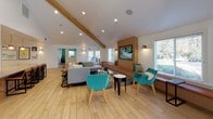 The Bella Vista Clubhouse is a thoughtfully designed single-story community gathering space that blends contemporary aesthetics with warm, inviting elements. The main lounge area serves as the heart of the clubhouse, featuring soaring vaulted ceilings accented by an exposed natural wood beam that adds architectural character. Light wood-tone flooring flows continuously throughout, enhancing the sense of openness and cohesion. Large windows invite abundant natural light, creating a bright and welcoming atmosphere. The lounge is furnished with a comfortable sectional sofa, vibrant teal accent chairs, and a built-in wood media wall with a large wall-mounted flat-screen TV and upholstered bench seating beneath — a perfect setting for community gatherings or relaxed socializing. A dedicated kitchenette provides practical amenities, featuring stone-look countertops, crisp white cabinetry with gold hardware, a stainless steel sink, textured tile backsplash, and pendant lighting overhead. A glass-enclosed leasing office with sliding glass doors offers a professional yet transparent workspace, complete with a stylish pendant light fixture and modern furnishings. Recessed lighting throughout ensures consistent illumination across all areas. The overall ambiance of the Bella Vista Clubhouse is modern, clean, and community-focused, making it an ideal hub for residents to connect and unwind.