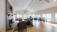 Located at 7820 W Capitol Ave in Little Rock, Arkansas, this single-story home is defined by its dramatic vaulted ceilings and open-concept layout that creates a generous, airy atmosphere throughout the main living area. Light hardwood flooring runs continuously across the space, complementing the neutral wall tones and contributing to a warm, welcoming ambiance.

The expansive living room serves as the centerpiece of the home, anchored by a painted brick fireplace adorned with a white mantle and a decorative sunburst mirror above. Multiple seating arrangements accommodate both relaxed lounging and conversation, while a ceiling fan overhead adds both function and character. Large windows dressed with white horizontal blinds line the walls, flooding the room with natural light and offering views of the outdoors. French doors at the rear provide direct access to the exterior.

Adjacent to the living area, a dedicated dining space features a dark wood table and chairs positioned beneath additional oversized windows. The kitchen connects to the main living space via a pass-through bar counter with granite-style surfaces and two bar stools, making it ideal for casual dining and entertaining. The kitchen area benefits from its own natural light source through a large side window. The thoughtful layout ensures seamless flow between the living, dining, and kitchen areas, making this home well-suited for both everyday living and gatherings.