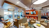 Located on North High Street in Columbus, Ohio, this dynamic community lounge space offers a generously sized, single-floor layout designed for social gathering and relaxation. The interior blends contemporary and eclectic design elements, anchored by a striking orange accent wall that frames a bar-style counter with white stools and dual wall-mounted televisions — an ideal hub for casual interaction. Distinctive pendant and drum-shade light fixtures hang from a coffered ceiling with warm wood accents, adding architectural depth throughout.

The lounge transitions seamlessly across distinct zones: a lively seating area furnished with an assortment of upholstered armchairs, leather sofas, and layered area rugs creates an inviting, relaxed atmosphere. A separate game area features a classic arcade cabinet alongside additional casual seating, adding a playful, recreational element. Bold "LUXE BELLE" typographic wall murals referencing the Columbus skyline serve as eye-catching focal points throughout the space.

Hexagonal patterned tile flooring contrasts beautifully with wide-plank wood-look flooring, defining different activity zones. Large floor-to-ceiling sliding glass doors flood the interior with natural light and provide direct access to an outdoor patio area, further expanding the usable social space. The overall ambiance is energetic yet comfortable, making it a well-appointed communal destination.