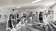 Located at 3301 Eamon Ct in Virginia Beach, VA, this fitness center offers a thoroughly equipped exercise facility designed to support a full range of workout routines. The single-floor space is generously sized, providing ample room for members to move freely between equipment zones without congestion.

The cardio area is outfitted with multiple treadmills, elliptical trainers, and additional cardiovascular machines, all positioned along windowed walls that allow natural light to fill the space. The large windows fitted with white blinds create a bright and energizing workout environment.

The strength training section is equally impressive, featuring a wide array of Cybex International and Eagle selectorized machines targeting various muscle groups, along with cable systems, adjustable benches, and a full rack of dumbbells organized along a mirrored wall. The floor-to-ceiling mirrors throughout the facility enhance the sense of openness and allow users to monitor their form during exercises.

Underfoot, durable dark rubber flooring provides safety and stability across the workout areas, transitioning to tile near the entrance. The textured ceiling is fitted with recessed fluorescent lighting panels that supplement the natural light. A front desk area with a mounted television monitor is present near the entrance. The overall ambiance is clean, bright, and professional, making it a well-maintained facility suited for comprehensive fitness training.