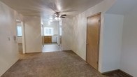 This single-story space offers a straightforward, functional layout with a seamlessly connected living area and kitchen. The living area is finished with wall-to-wall carpet in a neutral tan tone, providing a comfortable underfoot feel throughout. A ceiling fan with an integrated light fixture is centrally mounted on the textured ceiling, offering both ventilation and ambient lighting to the main area.

A wood-toned interior door is positioned along the right wall, providing access to an additional room or storage space. The open sightline from the living area leads directly into the kitchen, which features warm medium-toned wood cabinetry along the lower walls and what appears to be white appliances, including a dishwasher. A window above the kitchen area allows natural light to filter into the space, brightening the transition between rooms.

The walls throughout are finished in a neutral off-white tone, lending a clean, understated backdrop that complements the warm flooring and cabinetry. The overall ambiance is practical and modest, with a cohesive color palette that keeps the space feeling light and open.