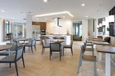 Located at 2204 Duluth Highway 120 in Duluth, Georgia, this single-floor community amenity space is designed with a clean, contemporary aesthetic that balances functionality with upscale style. The open-plan layout flows seamlessly from one zone to the next, anchored by light-toned herringbone hardwood flooring that adds warmth and visual interest throughout.

The fully equipped kitchen is a standout feature, showcasing rich walnut cabinetry paired with crisp white quartz countertops, stainless steel appliances including a French door refrigerator and double wall ovens, and a striking textured white accent wall. A large central island with a statement overhead range hood serves as a natural gathering point, with bar-height seating ideal for casual interaction.

Adjacent to the kitchen, the dining area offers multiple round tables and mixed seating arrangements bathed in natural light from expansive windows. Recessed and cove lighting throughout enhances the bright, airy atmosphere. A stylish lounge area provides comfortable seating arranged around a central coffee table, encouraging relaxed conversation.

A defined lounge zone along one wall features built-in banquette seating framed by sleek vertical black dividers, creating an intimate yet open atmosphere. Beyond, a fitness center is visible through glass partitions, reinforcing the space's commitment to a comprehensive amenity experience.