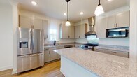 This thoughtfully designed single-floor home offers a clean, modern aesthetic throughout, unified by warm light hardwood flooring and crisp white walls. The open-concept living area is bright and generously proportioned, with multiple windows fitted with horizontal blinds that invite natural light and offer neighborhood views. A ceiling fan adds both style and comfort to the space.

The kitchen is a standout feature, showcasing an expansive granite-topped center island, shaker-style cabinetry in a soft greige finish, and a full suite of stainless steel appliances including a gas range, built-in microwave, dishwasher, and French door refrigerator. Pendant lighting and recessed fixtures illuminate the space, while a decorative tile backsplash and a stainless steel wall-mounted range hood add refined detail.

A clean, well-lit hallway with panel-style doors connects the rooms, providing a smooth flow throughout the home. The bathroom features a double-sink vanity with granite countertops, shaker cabinetry with brushed nickel hardware, a large mirror, and a walk-in shower with glass enclosure. An additional bathroom includes a combination tub and shower with brushed gold fixtures.

The attached garage offers a spacious concrete-floored area with a sectional overhead door, providing practical storage and parking. Overall, the home presents a fresh, move-in-ready environment with consistent finishes and functional design.