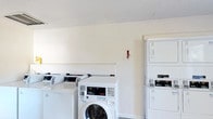 This single-floor laundry facility is designed with practicality and efficiency in mind. The room is outfitted with multiple commercial-grade top-loading and front-loading washing machines, as well as stacked dryer units, providing ample capacity for laundry needs. The machines are arranged in an organized manner, maximizing the use of the available floor space while maintaining clear pathways for ease of use. The interior features clean white walls and a textured ceiling that contribute to a bright, straightforward atmosphere. The room is well-lit, ensuring a functional environment at any time of day. Each machine appears to be equipped with digital controls and coin or card-operated payment systems, indicating a shared-use or communal laundry setup. A fire safety pull station is visibly mounted on the wall, reflecting attention to safety standards within the facility. The overall design prioritizes utility, with a simple and uncluttered layout that makes the space easy to navigate. This laundry room provides a convenient, no-frills solution for handling laundry needs efficiently.