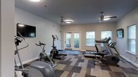 Located at 300 Autumn River Run in the Roxborough neighborhood of Philadelphia, PA, this single-floor fitness center offers a well-equipped and comfortable workout environment. The space features a thoughtfully designed layout with a variety of cardio machines, including stationary bikes, an elliptical, a rowing machine, and treadmills, providing residents with a comprehensive range of exercise options. A wall-mounted flat-screen television adds an entertainment element to workouts. The room is finished with a modern, patterned carpet in neutral tones of gray and beige, creating a clean and contemporary aesthetic. Two ceiling fans with integrated lighting ensure proper air circulation and comfort during exercise sessions. Natural light flows generously into the space through multiple windows fitted with blinds, as well as a set of glass-paneled double doors that open to a lush outdoor area, enhancing the overall ambiance. The walls are painted in a soft, neutral tone, contributing to an open and inviting atmosphere. This fitness center combines functionality and thoughtful design to support an active lifestyle.
