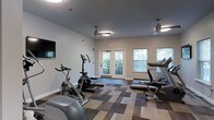Located at 300 Autumn River Run in the Roxborough neighborhood of Philadelphia, PA, this single-floor fitness center offers a well-equipped and comfortable workout environment. The space features a thoughtfully designed layout with a variety of cardio machines, including stationary bikes, an elliptical, a rowing machine, and treadmills, providing residents with a comprehensive range of exercise options. A wall-mounted flat-screen television adds an entertainment element to workouts. The room is finished with a modern, patterned carpet in neutral tones of gray and beige, creating a clean and contemporary aesthetic. Two ceiling fans with integrated lighting ensure proper air circulation and comfort during exercise sessions. Natural light flows generously into the space through multiple windows fitted with blinds, as well as a set of glass-paneled double doors that open to a lush outdoor area, enhancing the overall ambiance. The walls are painted in a soft, neutral tone, contributing to an open and inviting atmosphere. This fitness center combines functionality and thoughtful design to support an active lifestyle.