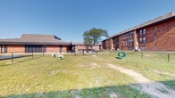 Located at 6300 Century City S in Reynoldsburg, Ohio, this outdoor courtyard space offers an open, functional area set within a well-established brick building complex. The surrounding structures are constructed of traditional red brick with large windows and low-pitched rooflines, lending the environment a grounded, institutional character that speaks to durable, practical architecture.

The courtyard itself is a grassy, open-air area framed on multiple sides by the adjacent buildings, creating a naturally enclosed and sheltered atmosphere. A mature tree stands prominently near the center background, providing a focal point and a sense of established greenery. The grounds feature light play or recreational elements, including animal-shaped figures and a circular green structure, suggesting this space is designed to accommodate active, outdoor use.

A perimeter fence delineates the boundaries of the space, adding a layer of structure and definition to the area. Beneath a clear, open sky, the courtyard benefits from abundant natural light throughout the day, enhancing the sense of openness. The combination of surrounding multi-story buildings, mature landscaping, and dedicated recreational features makes this an informative and purposeful outdoor environment suited for community or recreational activity.