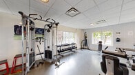 This fitness center is a fully equipped, single-floor facility designed to support a wide range of workout routines. Upon entry, visitors pass through a carpeted entryway with decorative frosted glass doors featuring a city map motif, leading into the main exercise areas. Restroom facilities are conveniently located near the entrance.

The main workout floor is finished with durable hard flooring and features a row of modern cardio machines, including treadmills and ellipticals, positioned along windowed walls that invite natural light and views of the surrounding greenery. A dedicated free-weight area offers a full dumbbell rack alongside a multi-functional cable machine, providing options for both isolation and compound exercises. Additional strength training machines are positioned throughout the space to accommodate a variety of muscle groups.

A standout feature is the full wall of floor-to-ceiling mirrors, which visually expands the space while supporting form checks during workouts. A ballet barre runs along the mirror wall, indicating use for dance or flexibility training. The facility also includes a heavy punching bag, stability balls, and step platforms for functional fitness. The drop ceiling with recessed lighting provides consistent, even illumination throughout. The overall ambiance is clean, practical, and well-organized, offering a professional environment for fitness training.