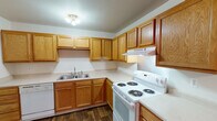 Located at 3510 N Drake Rd in Kalamazoo, MI, this single-floor unit offers a practical and functional layout throughout. The kitchen is arranged in an L-shaped configuration and features warm oak cabinetry with ample upper and lower storage, light-toned countertops, a double-basin stainless steel sink, a white electric coil range, and a dishwasher. Dark hardwood-style flooring adds contrast to the kitchen's neutral palette. The living and dining area transitions seamlessly from hardwood-style flooring to plush carpeting, with a ceiling fan overhead and a large window fitted with vertical blinds that draws in natural light. The bedrooms are carpeted and finished with neutral walls, offering a calm and comfortable atmosphere. One bedroom includes a built-in closet with sliding wood-paneled doors, while another features a window overlooking mature outdoor trees. The unit includes two bathrooms: a full bath with a tiled tub-and-shower combination, tile flooring, and a wood-framed vanity, as well as a half bath with tile flooring, a wall-mounted mirror, and a grab bar. The overall design is straightforward and well-maintained, providing a clean and move-in-ready environment.