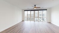 Located at 945 Mid City Drive in Huntsville, Alabama, this thoughtfully designed unit offers a clean, modern aesthetic across a single floor. The open-concept kitchen and living room serve as the heart of the space, where warm hardwood-style flooring and crisp white walls create a bright, welcoming atmosphere. The living area is anchored by floor-to-ceiling windows that span the exterior wall, flooding the space with natural light and offering views of the surrounding mid-city development. A ceiling fan adds both style and function overhead. The kitchen is well-appointed with light wood flat-front cabinetry, stainless steel appliances including a French door refrigerator, electric range, built-in microwave, and dishwasher. A tiled backsplash with a subtle mosaic pattern, light quartz countertops, a deep undermount sink with a matte black faucet, and open industrial-style shelving complete the culinary space. Pendant lighting adds a modern touch above the counters. The bathroom features a frameless glass walk-in shower, large-format tile flooring, and a wood-accented vanity with a stone countertop, maintaining the unit's clean, contemporary character. A dedicated closet provides practical storage. The overall design is cohesive and modern, ideal for efficient urban living.
