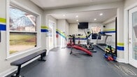 Located at 524 Bramlett Rd in Charlotte, NC, this compact yet efficiently designed fitness facility offers a complete workout experience within a single-floor layout. The exercise room serves as the centerpiece of the space, boasting durable dark rubber flooring ideal for heavy equipment use. A vibrant decorative accent — bold horizontal stripes in blue, green, and yellow — runs along the walls, lending the room an energetic and motivating atmosphere. Recessed lighting throughout ensures the space is well-lit, while multiple windows along the walls invite natural light and provide views of the surrounding outdoor greenery. The exercise room is equipped with a multi-station cable machine, a treadmill, an elliptical trainer, a stationary spin bike, an adjustable workout bench, free weights, and a wall-mounted flat-screen television for entertainment during workouts. A large mirror along one wall enhances the sense of space and supports proper form. The attached bathroom is clean and functional, featuring tile flooring, a vanity with white cabinetry, and all essential fixtures. Together, these two rooms create a self-contained, purpose-built fitness environment that is both practical and visually dynamic.
