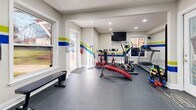 Located at 524 Bramlett Rd in Charlotte, NC, this compact yet efficiently designed fitness facility offers a complete workout experience within a single-floor layout. The exercise room serves as the centerpiece of the space, boasting durable dark rubber flooring ideal for heavy equipment use. A vibrant decorative accent — bold horizontal stripes in blue, green, and yellow — runs along the walls, lending the room an energetic and motivating atmosphere. Recessed lighting throughout ensures the space is well-lit, while multiple windows along the walls invite natural light and provide views of the surrounding outdoor greenery. The exercise room is equipped with a multi-station cable machine, a treadmill, an elliptical trainer, a stationary spin bike, an adjustable workout bench, free weights, and a wall-mounted flat-screen television for entertainment during workouts. A large mirror along one wall enhances the sense of space and supports proper form. The attached bathroom is clean and functional, featuring tile flooring, a vanity with white cabinetry, and all essential fixtures. Together, these two rooms create a self-contained, purpose-built fitness environment that is both practical and visually dynamic.