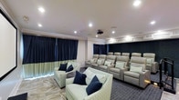 This single-floor home theater is thoughtfully designed to deliver a true cinema experience. The room is arranged with tiered seating to ensure clear sightlines, featuring plush light gray leather recliner chairs equipped with armrests and cup holders, positioned in stadium-style rows toward the rear. The front area of the room accommodates additional casual seating in the form of patterned fabric armchairs accented with navy and green throw pillows, offering a relaxed viewing alternative.

A ceiling-mounted projector serves as the primary display source, directed toward the front of the room. Recessed lighting provides ambient illumination, while dark navy blackout curtains line the walls, effectively blocking external light and enhancing the viewing environment. Acoustic wall panels are incorporated into the design to optimize sound quality within the space.

The flooring transitions from patterned tile near the entrance to dark carpet in the seating area, subtly defining zones within the room. Crown molding and clean, neutral wall tones contribute to a polished, finished aesthetic. Overall, this theater room balances functional design with comfortable, upscale furnishings.