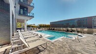 Located at 4810 Bradford Dr NW in Huntsville, Alabama, this outdoor amenity space presents a well-appointed resort-style pool deck nestled within a contemporary multi-story building complex. The centerpiece is a generously sized swimming pool with a striking geometric tile pattern visible through the crystal-clear water, complemented by a separate spa for relaxation. The pool is framed by a broad paved deck finished in patterned brick and stone pavers, offering ample room for leisure and socializing.

Rows of modern white lounge chairs line both sides of the pool, providing abundant sunbathing opportunities, while shaded seating areas with umbrellas offer respite from the sun. An additional outdoor patio area extends alongside the building, furnished with lounge chairs, fire pit-style tables, umbrella seating, and outdoor grilling stations — ideal for gatherings and al fresco entertaining. String lights overhead add ambiance to the terrace area.

The space is enclosed on multiple sides by the architecturally distinct multi-story building, which features a mix of brick cladding, large windows, and private balconies overlooking the pool deck. Open views toward the surrounding Huntsville cityscape and tree canopy are visible beyond the perimeter, enhancing the outdoor experience with natural scenery and an open-sky atmosphere.