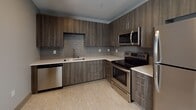 Located at 183 E Main St in the heart of downtown Rochester, NY, this modern single-floor residence offers a thoughtfully designed layout with contemporary finishes throughout. The kitchen is a standout feature, showcasing dark wood-grain cabinetry that extends floor to ceiling, light quartz countertops, and a gray subway tile backsplash. A full suite of stainless steel appliances — including a refrigerator, electric range, over-the-range microwave, and dishwasher — completes the space, combining style with full functionality. Light vinyl plank flooring runs through the kitchen and living areas, adding warmth and cohesion to the open layout. The main living area is bright and open, with clean lines, neutral-toned walls, and recessed architectural ceiling details that enhance the sense of space. A louvered closet door and dedicated utility area with a stacked washer and dryer add convenient in-unit storage and laundry access. The bathroom is finished to a high standard, featuring a dark wood vanity with quartz countertop, a large framed mirror, and a walk-in glass-enclosed shower with built-in shelving and chrome fixtures. The overall aesthetic is clean, modern, and move-in ready, making this a well-appointed residence in a prime urban location.