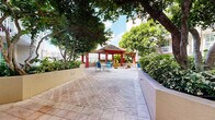 Located at 410 Evernia Street in the heart of West Palm Beach, Florida, this outdoor courtyard space offers a well-designed, single-level retreat surrounded by tropical landscaping. The patio is finished with a classic herringbone-pattern brick paver surface that extends throughout the space, providing a clean and durable foundation underfoot.

A standout architectural feature is the red-columned gazebo with a pitched, tile-style roof, positioned toward the waterfront edge of the courtyard. It provides shaded seating and serves as a focal point within the layout. Multiple seating areas are distributed across the patio, furnished with glass-top tables and blue sling-style chairs, accommodating both small and larger gatherings.

Mature trees with broad canopies are planted in raised stucco planters, casting natural shade and creating a lush, garden-like atmosphere. Tropical shrubs and colorful flowering plants line the perimeter, enhancing the sense of enclosure and privacy. A small decorative gravel garden with a birdbath adds a tranquil accent near the gazebo.

The courtyard is framed by a multi-story building with balconies and large windows, whose ground-floor interior spaces are accessible directly from the patio through glass doors. The overall ambiance is bright, open, and inviting, reflecting the relaxed outdoor lifestyle characteristic of South Florida.