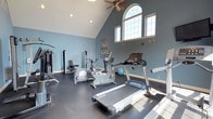 Located at 105 Amberley Dr in Blue Bell, PA, this single-floor fitness center is thoughtfully designed to support a well-rounded workout experience. The space is defined by striking vaulted ceilings that soar overhead, creating an open and airy atmosphere enhanced by soft blue-gray walls that lend a calm, focused ambiance. Natural light pours in through multiple windows, including a prominent arched transom window that serves as an architectural highlight, brightening the room throughout the day. Ceiling fans help maintain comfortable airflow during workouts. The flooring features durable, dark rubberized material well-suited for fitness use. The room is equipped with a comprehensive selection of cardio machines, including treadmills, elliptical trainers, and a stationary bike, arranged to allow easy movement throughout the space. A multi-function cable and strength training machine anchors one side of the room, offering versatile resistance exercise options. A wall-mounted television provides entertainment during workouts. Framed artwork and a stability ball add finishing touches that make the space feel both functional and welcoming. Overall, this fitness center combines practical amenities with an inviting design to offer an effective and comfortable environment for exercise.