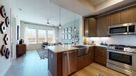 This well-designed one-bedroom, one-bathroom unit offers a thoughtfully planned single-level layout. The open-concept kitchen is a standout feature, showcasing rich walnut-toned cabinetry, quartz countertops, a farmhouse-style stainless steel sink, and a full suite of stainless steel appliances including a gas range, microwave, refrigerator, and dishwasher. Geometric pendant lights hang above the kitchen island, which provides bar seating and serves as a natural divider between the kitchen and living area. Light wood-style flooring flows throughout the main living spaces, enhancing the bright, modern atmosphere. The living room benefits from large windows that invite abundant natural light and offer outdoor views, with a ceiling fan adding functional comfort. A dedicated workspace nook is integrated seamlessly into the living area. The bedroom features plush carpeting, dual windows with outdoor views, and soft neutral tones that create a calm retreat. The full bathroom includes a tub-shower combination with tiled surround, dark cabinetry with gold-toned hardware, and light tile flooring, delivering a clean, contemporary finish throughout.