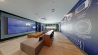 Located in Shoreline, WA, this single-floor Game Zone lounge is a dedicated entertainment and social space thoughtfully designed around a sports-viewing experience. The room centers on a striking large-format video wall composed of multiple screens, flanked on both sides by dark built-in shelving units displaying an assortment of sports memorabilia, including footballs, basketballs, and other collectibles. Recessed lighting and a ceiling-mounted HVAC unit keep the space comfortable and well-lit throughout. The seating area is anchored by a generously sized sectional sofa in warm taupe tones, complemented by a bold cognac leather ottoman that serves as a central gathering point. Light hardwood flooring and a neutral area rug add warmth and texture underfoot. A long bar-height communal table with dark chairs occupies the center of the room, offering an alternative perch for watching the action. One wall features an illuminated "Game Zone" sign set against a richly textured moss-panel backdrop in green and gold tones, while an adjacent wall showcases a bold blue mural depicting architectural blueprints of NFL stadiums across the National Football Conference. Together, these elements create an immersive, energetic ambiance purpose-built for sports fans and community entertainment.