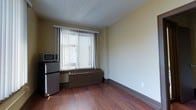 This single-floor space offers a straightforward and functional layout designed for efficient living. The room features warm hardwood flooring that runs throughout, complementing the neutral, light-toned walls that create a clean and understated atmosphere. A large window fitted with vertical blinds serves as the focal point of the main area, allowing natural light to filter in while offering privacy control. Beneath the window sits a built-in HVAC unit, ensuring year-round climate comfort. To one side, a compact appliance station includes a stainless steel mini-refrigerator and a countertop microwave, providing essential kitchen conveniences without requiring a dedicated kitchen space. The white flat ceiling and wide baseboards contribute to a tidy, well-finished interior. The overall ambiance is modest and practical, suited for straightforward day-to-day use. The space is move-in ready with core utilities and appliances already in place, making it a functional option for those seeking a no-frills, self-contained living or working environment.