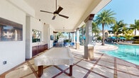 This covered outdoor amenity space offers a resort-style experience designed for relaxation and recreation. The structure features a pitched, textured ceiling supported by sturdy square columns, creating a shaded pavilion that seamlessly blends indoor comfort with outdoor living. Ceiling fans are mounted overhead to ensure air circulation, keeping the space comfortable in warm weather.

A prominent game table—covered and positioned centrally—serves as a focal point of the space, inviting leisurely activity. Along one wall, rich dark-wood cabinetry with a built-in wet bar and sink provides convenient access to refreshments, making this area well-suited for entertaining.

The flooring features a decorative tile pattern in warm earth tones, adding visual interest underfoot. Open on multiple sides, the pavilion offers unobstructed views of a sparkling pool and lush landscaping filled with tall palm trees, enhancing the tropical ambiance. A wall-mounted flat-screen television is also present, adding a modern entertainment element.

A separate lounge area with blue seating is visible beyond the columns, further extending the space's functionality. The overall atmosphere is bright, airy, and inviting, with abundant natural light filtering through the open sides and evoking the feel of a luxury resort retreat.