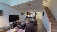 This two-floor space presents an open-concept layout on the main level, where the living area flows directly into the kitchen. The living area is furnished with a plush dark sectional sofa, a swivel accent chair, and a leather storage ottoman, all anchored by a bold, multicolored geometric area rug that adds a lively visual focal point to the room. A large flat-screen television is mounted on a low media console along one wall, complemented by framed artwork that adds character to the space.

The kitchen is positioned at the rear of the main level and features rich dark wood cabinetry, a white refrigerator, and a raised breakfast bar with sleek modern bar stools. The compact, efficient layout makes good use of the available space. Hardwood-style flooring runs through the kitchen and hallway areas, providing a clean contrast to the carpeted living zone.

A hallway extends from the main living area, leading to additional rooms and a staircase connecting to the second floor. The ceilings are standard height, with recessed lighting and visible HVAC vents providing a functional, well-maintained environment throughout.