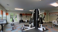 This single-floor fitness room offers a functional and practical workout environment designed to accommodate a variety of exercise needs. The space is outfitted with a multi-station cable and weight machine as a central strength-training fixture, complemented by cardio equipment including an elliptical trainer and a stationary cycle. A wall-mounted display screen adds a modern touch for entertainment or guided workouts during cardio sessions. The flooring features a multi-colored rubber tile layout, providing cushioning and durability suited for gym use. A full-length mirror along one wall enhances the sense of space while allowing users to monitor their form. The walls are accented with a warm terracotta-toned horizontal band contrasting against neutral grey tones, giving the room a distinct, energized visual character. Overhead fluorescent lighting ensures consistent illumination throughout. A set of glass-paned double doors at the far end of the room floods the space with natural light and offers views of lush greenery outside, contributing to an open and inviting atmosphere. The overall layout is organized and accessible, making it a convenient facility for daily fitness routines.