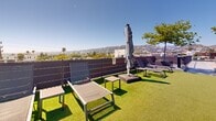 Located at 801 N Fairfax Ave in the heart of West Hollywood, this expansive rooftop deck offers a thoughtfully designed outdoor retreat with a variety of distinct zones for relaxation and social gathering. The space is defined by its clean, contemporary aesthetic, blending warm wood decking with polished concrete surfaces and dark metal accents throughout.

At one end, a built-in outdoor kitchen features stainless steel grills and ample counter space, ideal for al fresco dining. String lights overhead add a welcoming ambiance during evening gatherings. A covered pergola with a sleek canopy provides shaded seating, bordered by mature trees planted in oversized modern planters alongside decorative gravel beds that lend a sophisticated, low-maintenance landscape design.

A dedicated turf lounge area offers chaise loungers and side tables beneath a retractable umbrella, perfectly positioned to take in the panoramic views of the Hollywood Hills and the broader Los Angeles cityscape. Manicured hedges in large planters and horizontal wood privacy screens define the boundaries while maintaining an open, airy feel.

The rooftop's layout, as revealed in an aerial perspective, stretches across a generous footprint with multiple gathering nodes connected by wide walkways, making it well-suited for both quiet relaxation and larger social events under the iconic Southern California sun.