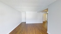 This single-floor space presents a clean, minimalist aesthetic throughout. The main living area is immediately welcoming, characterized by rich, warm-toned hardwood floors that extend seamlessly across the open layout, creating a sense of continuity and flow. Crisp white walls reflect light effectively, contributing to the bright and airy atmosphere of the space. The textured popcorn ceiling adds a classic touch overhead, while white baseboards provide a clean contrast to the hardwood flooring below. A partial interior wall divides areas within the open floor plan, subtly defining separate zones without sacrificing the overall openness. A doorway visible in the background leads to additional rooms, hinting at a well-connected layout. To the right, a hallway with a ceiling fan leads further into the space, suggesting access to private rooms or a bathroom, with natural light filtering through from adjacent areas. The overall ambiance is neutral and versatile, offering a blank canvas well-suited for a variety of interior styles. The consistent use of white throughout the walls and trim reinforces a cohesive, tidy appearance across the entire floor.