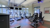 This fitness center is a thoughtfully designed, single-floor workout facility that offers a comprehensive range of exercise equipment in a bright and welcoming environment. The space is outfitted with multiple treadmills, elliptical trainers, and a stationary bike, providing ample options for cardiovascular training. Strength and resistance equipment is also available, catering to a full-body workout experience.

Large windows line the walls, flooding the interior with abundant natural light and offering pleasant views of the lush, tree-lined surroundings outside. The flooring features a durable, multi-toned rubber surface in a checkered pattern, combining both functionality and visual appeal. Overhead, ceiling fans help maintain comfortable airflow throughout the space, while modern track lighting ensures consistent illumination during all hours.

Multiple flat-screen televisions are mounted at eye level along the walls, allowing users to stay entertained during their workouts. The neutral-toned walls and open layout contribute to a clean, airy ambiance. Overall, this fitness center is designed to be both practical and comfortable, making it a well-rounded facility suited for a variety of fitness routines.