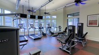 This fitness center is a thoughtfully designed, single-floor workout facility that offers a comprehensive range of exercise equipment in a bright and welcoming environment. The space is outfitted with multiple treadmills, elliptical trainers, and a stationary bike, providing ample options for cardiovascular training. Strength and resistance equipment is also available, catering to a full-body workout experience.

Large windows line the walls, flooding the interior with abundant natural light and offering pleasant views of the lush, tree-lined surroundings outside. The flooring features a durable, multi-toned rubber surface in a checkered pattern, combining both functionality and visual appeal. Overhead, ceiling fans help maintain comfortable airflow throughout the space, while modern track lighting ensures consistent illumination during all hours.

Multiple flat-screen televisions are mounted at eye level along the walls, allowing users to stay entertained during their workouts. The neutral-toned walls and open layout contribute to a clean, airy ambiance. Overall, this fitness center is designed to be both practical and comfortable, making it a well-rounded facility suited for a variety of fitness routines.