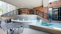 This single-level covered amenity space at Kings Landing offers a well-appointed retreat designed for relaxation and leisure. The focal point of the area is a tiered water feature, prominently displaying the Kings Landing branding, with small jets creating a gentle, flowing fountain effect. Adjacent to the fountain sits a built-in hot tub, finished in neutral stone tiling that complements the surrounding hardscape. The space is enclosed by warm exposed brick walls that lend a classic, grounded aesthetic, while sleek black wrought iron railings line a staircase leading to an upper landing area. Above, a structured ceiling with recessed grid panels provides overhead coverage, keeping the space sheltered while maintaining an open, airy feel. A wooden bench and ornamental iron seating add functional yet decorative touches throughout. The combination of masonry, metalwork, and water elements creates a cohesive, resort-inspired ambiance suited for both casual lounging and unwinding after a long day.
