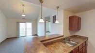 This single-floor space showcases an open-concept layout that seamlessly connects the kitchen and living area, creating a spacious and functional environment. The kitchen features warm-toned granite-look countertops that extend into a peninsula, providing both additional workspace and a casual dining or entertaining surface. A stainless steel sink sits along the counter, complemented by rich medium-toned wood cabinetry. Three elegant pendant lights hang above the kitchen area, drawing the eye upward to the impressively vaulted ceilings, which amplify the sense of openness throughout the space. The living area is finished with carpet flooring and benefits from generous natural light streaming in through sliding glass doors that open to a private balcony with a metal railing, offering outdoor access and views. A doorway visible from the main living area leads to an additional room, suggesting a well-connected floor plan. The overall ambiance is bright, airy, and welcoming, with the combination of vaulted ceilings, natural light, and an open layout making the space feel larger than its footprint suggests.