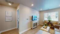 This single-floor residence is thoughtfully designed with a cohesive neutral palette and modern finishes that create a clean, welcoming atmosphere. The layout, as seen in the overhead view, flows efficiently between a living area, kitchen, bedroom, and bathroom.

The living area features light wood-look flooring, recessed lighting, and a window with blinds that allows natural light to brighten the space. A white glass-door media cabinet provides functional storage beneath a wall-mounted television, while a sitting area with a sofa, ottoman, and a small bistro table near the window offers versatile use of the room.

The kitchen is well-equipped with white shaker-style cabinetry, light quartz countertops, and stainless steel appliances including a refrigerator, range, and dishwasher. Matte black hardware and a matching black faucet add a contemporary contrast against the bright cabinetry and wood-look flooring.

The bedroom features plush carpeting, a large window with natural light overlooking the exterior, and ample space for a full furniture arrangement including a dresser and nightstands.

The bathroom includes a tub-shower combination with white subway tile, a single vanity with quartz countertop, white cabinetry, and matte black fixtures that tie the design together cohesively.
