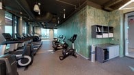 Located at 2 I St SE in Washington, DC, this contemporary fitness center is designed to deliver a well-equipped and visually stimulating workout environment. The space is organized across a single floor, with distinct zones dedicated to various types of exercise. The cardio area is generously outfitted with a full lineup of treadmills, elliptical machines, stair climbers, and stationary bikes, including Life Fitness cycling bikes arranged alongside a striking textured feature wall finished in weathered green tones that adds an industrial-artistic character to the space. A dedicated free weight and functional training area provides additional workout versatility, complete with stability balls and storage. Throughout, the flooring is cushioned rubber, ideal for high-impact activity. The ceiling is exposed, showcasing ductwork and modern geometric pendant lighting fixtures that illuminate the space with a clean, bright ambiance. Expansive floor-to-ceiling windows flood the interior with natural light and frame sweeping views of the Washington, DC skyline, creating an inspiring backdrop for workouts. A water station with a bottle-filling dispenser and towel service station are conveniently positioned within the space. The overall design balances urban industrial aesthetics with functional fitness amenities.