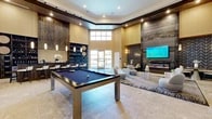 Located at 8901 Holly Ave N in Albuquerque, NM, this upscale clubroom is a single-floor amenity space designed with a modern, resort-inspired aesthetic. Soaring double-height ceilings with a recessed tray detail and abundant natural light from oversized windows create an open, airy atmosphere throughout.

The space is anchored by a sleek billiards table positioned on large-format travertine tile flooring, serving as the room's central focal point. Adjacent to it, a fully appointed wet bar features floor-to-ceiling shelving stocked with glassware and bottles, pendant lighting, and a row of bar-height seating — ideal for social gatherings.

A dedicated lounge area is furnished with contemporary rounded swivel chairs, soft ottomans, and layered area rugs, fostering an inviting environment for conversation and relaxation. Opposite the lounge, a dramatic feature wall showcases a wide-format linear fireplace beneath a large wall-mounted television, flanked by textured chevron tile accents and warm sconce lighting.

Architectural details include wood-paneled accent walls, fine textured wallcovering, and decorative botanical wall art that add warmth and visual interest. Glass-paned French doors connect to adjacent spaces, enhancing the flow and openness of the overall layout. The combination of refined materials and thoughtful design makes this clubroom a standout gathering space.
