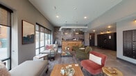 Located at 122 Parsons Ave in Columbus, Ohio's 43215 zip code, this single-floor space serves as the lobby and common area for Yardley, offering a distinctive and thoughtfully designed environment. The centerpiece of the space is a striking floor-to-ceiling illustrated mural wall that pays homage to the "Olde Towne East" neighborhood, setting a rich, locally inspired tone throughout. A dedicated reception desk positioned in front of the mural provides a clear focal point and welcoming arrival experience. The lounge area is furnished with an eclectic mix of seating, including a neutral sectional sofa, a rich olive-green armchair, and a warm terracotta accent chair, all arranged around natural wood coffee tables to create an inviting, social atmosphere. Recessed lighting and pendant fixtures illuminate the space, complementing the exposed ductwork detail that adds an industrial character to the otherwise warm interior. Large floor-to-ceiling windows along one wall flood the space with natural light and provide a visual connection to the exterior. Additional building amenities, including what appears to be a secure package locker system, are integrated along the perimeter. The overall ambiance balances modern industrial elements with cozy, residential warmth, making it a functional and visually engaging communal space.