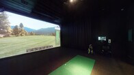 This single-level indoor golf simulator space is purpose-built for an immersive practice and playing experience. The room is finished entirely in matte black — walls, ceiling, and flooring — creating a darkened, distraction-free environment that maximizes the visual impact of the large projection screen dominating the forward wall. The screen displays full course simulations, complete with real-time shot data including carry distance, ball speed, and launch metrics, making it suitable for both casual play and serious performance tracking.

The hitting area centers on a dual-tone synthetic turf mat, designed to replicate fairway and tee conditions underfoot. The dark rubberized flooring surrounding the mat provides a stable, low-reflectance surface throughout the bay. A dedicated club stand keeps a full set of golf clubs organized and accessible alongside the hitting station. A compact control station with a monitor is positioned to the side for easy access during sessions.

To one side of the room, a small seating area with a wooden stool and table provides a comfortable spot for observers or players waiting their turn. Heavy black drapes line the walls, serving both as acoustic dampening and to contain the visual focus of the space. The room's layout is efficient and well-considered, delivering a fully functional simulator bay with a clean, professional aesthetic.