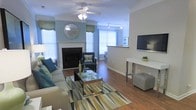 This single-floor space offers a thoughtfully designed interior that blends comfort and style seamlessly. The living area serves as the centerpiece, showcasing warm hardwood-style flooring that extends throughout the open-concept layout. A classic white-mantled fireplace anchors one wall, adding both a focal point and a cozy ambiance to the room. The walls are accented with a cool blue tone, complemented by crisp white crown molding and trim that lend an elegant, polished finish. Natural light pours in through multiple windows dressed with decorative valances and blinds, creating a bright and welcoming atmosphere. A ceiling fan with integrated lighting ensures year-round comfort. The living space is well-proportioned, accommodating a comfortable seating arrangement around a glass-top coffee table atop a bold geometric area rug. A sleek media console and wall-mounted television provide modern entertainment functionality. Track lighting along the ceiling offers additional illumination, while a glimpse into an adjoining area suggests a connected kitchen or dining space, enhancing the overall sense of flow and openness throughout this inviting home.