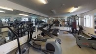 Located at 5001 College St SE in Lacey, Washington, this fitness center offers a comprehensive workout environment housed within a single-floor layout. The space features a textured ceiling with flush fluorescent lighting that provides bright, even illumination throughout. Full-length mirrors line the walls, creating a sense of expanded space and allowing users to monitor their form during exercise.

The facility is well-equipped with a broad selection of cardio machines, including treadmills, elliptical trainers, stair climbers, and rowing machines, arranged to maximize floor space and ease of movement. A dedicated strength training area includes multi-station cable machines, free weight benches, barbells, and a variety of weight plates, accommodating both beginner and experienced lifters.

Carpeted flooring runs throughout the space, providing a practical and comfortable surface underfoot. Natural light filters in through windows along one wall, supplementing the interior lighting and offering views of the surrounding outdoor environment. Wall-mounted sanitation dispensers are conveniently placed near the entrance, promoting cleanliness and hygiene. A scale is also available for users. The overall layout is functional and organized, making efficient use of the available floor space to support a wide range of fitness activities.