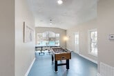 Located at 248 Debuys Rd in Biloxi, Mississippi, this single-room game space is purpose-built for recreation and entertainment. The room features a striking blue epoxy-style floor that sets a lively, energetic tone throughout. Vaulted ceilings rise overhead, lending an open, airy feel that makes the space feel larger than its compact footprint suggests. A textured ceiling with ceiling fans provides comfortable airflow, while multiple windows—including a prominent arched window at the front—flood the room with natural light and offer views of the outdoor surroundings. The game room is well-equipped with a full-size ping-pong table and a foosball table, both positioned to allow comfortable play. Wall art, including a "Table Tennis Life Lessons" poster, adds personality and a playful thematic touch. A secondary alcove area provides additional open floor space, accented by a floor lamp and wall-mounted decor including a tic-tac-toe game, offering flexibility for other activities. A dedicated exterior door provides convenient direct access, making this game room a self-contained entertainment destination that's as functional as it is fun.