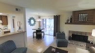 This single-floor residence offers an open and functional layout that seamlessly connects the living and dining areas into one cohesive space. The living room is anchored by a classic brick fireplace with a dark wood mantel, providing a warm and inviting focal point. A textured ceiling adds character overhead, while light hardwood flooring extends throughout, lending warmth and continuity to the space. The dining area is defined by a stylish pendant chandelier and a bistro-style dining set, complemented by a decorative sunburst mirror that adds an artistic touch to the wall. Large sliding glass doors flood the interior with natural light and offer a direct view to the outdoors, creating a bright and airy atmosphere. Dark curtain panels frame the glass doors, providing contrast and privacy when needed. The living area accommodates comfortable seating and flows naturally toward the dining space, making the layout ideal for both everyday living and entertaining. Neutral wall tones throughout keep the space feeling clean and versatile, while carefully placed décor accents add personality without overwhelming the room. Overall, the space presents a balanced blend of comfort, style, and practicality.