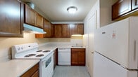 This single-floor, one-bedroom, one-bathroom unit offers a practical and efficient layout across a compact but thoughtfully arranged floor plan. The living room serves as the central gathering space, featuring neutral-toned carpet, a ceiling fan with light fixture, and a sliding glass door that opens to an outdoor patio area enclosed by a lattice screen. A side window allows additional natural light to enter the room, creating a bright and airy atmosphere.

The kitchen is arranged in an L-shaped configuration, equipped with warm-toned wood cabinetry extending to the upper walls for ample storage. White appliances, including an electric coil range, dishwasher, and refrigerator, are provided alongside a double basin sink and light-toned countertops. The flooring in the kitchen features a tile-style surface, offering easy maintenance.

The bedroom is a cozy, carpeted room with a window overlooking greenery and a sliding-door closet providing organized storage space. A secondary door in the bedroom provides additional access.

The bathroom includes a single-sink vanity with warm wood cabinetry, a tub-shower combination with tiled surround, a toilet, and tile-style flooring. A window above the tub provides natural light and ventilation. A hallway connects all rooms, completing the efficient and functional layout of this unit.